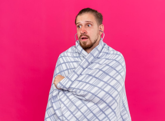 Free photo impressed young handsome slavic ill man wearing stethoscope wrapped in plaid looking at side isolated on pink wall with copy space