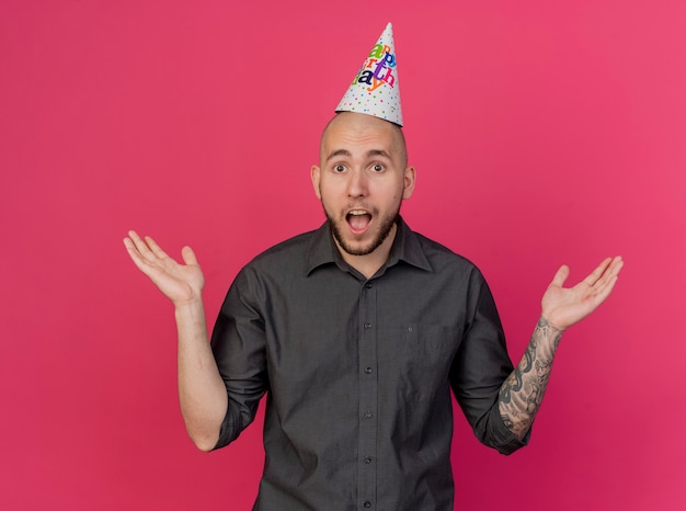 Impressed young handsome slavic party guy wearing birthday hat looking at camera showing empty hands isolated on crimson background