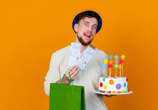 Impressed young handsome slavic party guy wearing party hat holding airplane tickets paper bag and birthday cake with stars looking at camera isolated on orange background with copy space