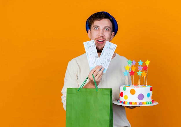 Impressed young handsome slavic party guy wearing party hat holding airplane tickets paper bag and birthday cake with stars looking at camera isolated on orange background with copy space