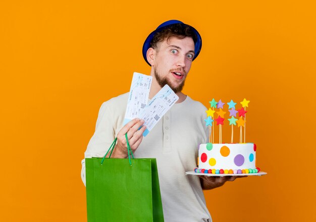 Impressed young handsome slavic party guy wearing party hat holding airplane tickets paper bag and birthday cake with stars looking at camera isolated on orange background with copy space