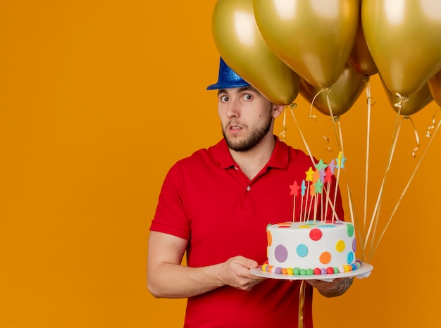 Impressed young handsome slavic party guy wearing party hat holding balloons and birthday cake looking at camera isolated on orange background with copy space