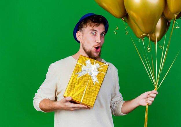 Foto gratuita impressionato giovane ragazzo slavo bello del partito che indossa il cappello del partito che tiene palloncini e confezione regalo che guarda l'obbiettivo isolato su priorità bassa verde