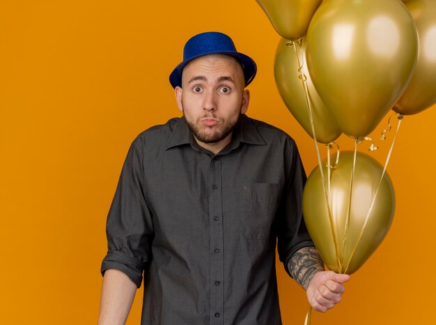 Impressed young handsome slavic party guy wearing party hat holding balloons looking at camera isolated on orange background with copy space