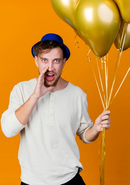 Foto gratuita colpito il giovane ragazzo slavo bello del partito che indossa il cappello del partito che tiene i palloncini che esaminano bisbigliando davanti isolato sulla parete arancione