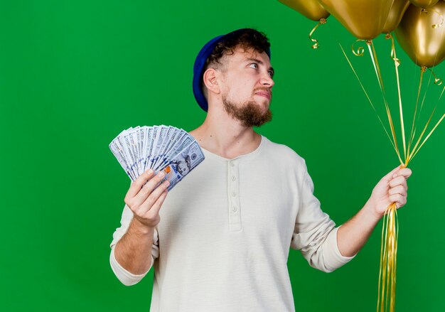 Impressed young handsome slavic party guy wearing party hat holding balloons and money isolated on green background with copy space