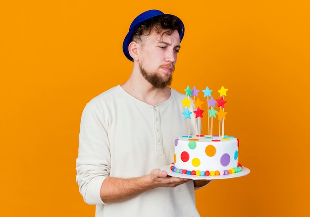 Impressed young handsome slavic party guy wearing party hat holding birthday cake with stars looking straight isolated on orange background with copy space