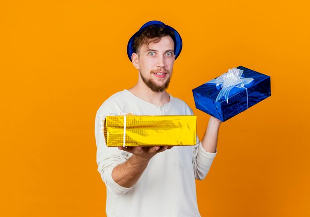 Impressed young handsome slavic party guy wearing party hat holding gift boxes stretching out one of them and looking at camera isolated on orange background with copy space