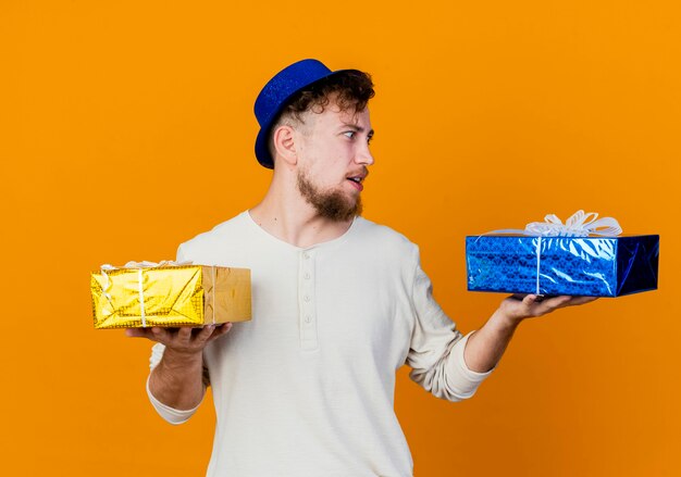 Impressed young handsome slavic party guy wearing party hat holding and looking at gift boxes isolated on orange background
