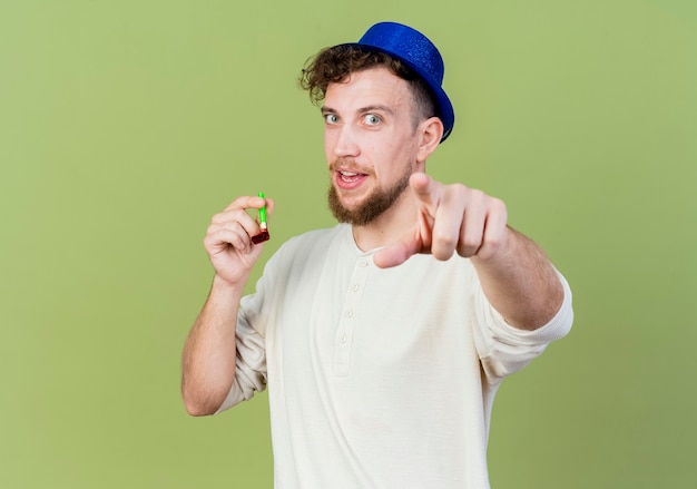 Impressed young handsome slavic party guy wearing party hat holding party blower looking and pointing at camera isolated on olive green background with copy space