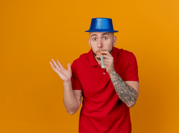 Impressed young handsome slavic party guy wearing party hat looking at camera showing empty hand blowing party blower isolated on orange background with copy space