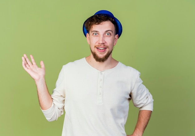 Impressionato giovane ragazzo slavo bello del partito che indossa il cappello del partito che guarda l'obbiettivo che mostra la mano vuota che tiene un altro sulla vita isolata su fondo verde oliva con lo spazio della copia