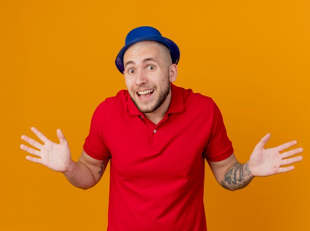 Impressed young handsome slavic party guy wearing party hat looking at camera showing empty hands isolated on orange background