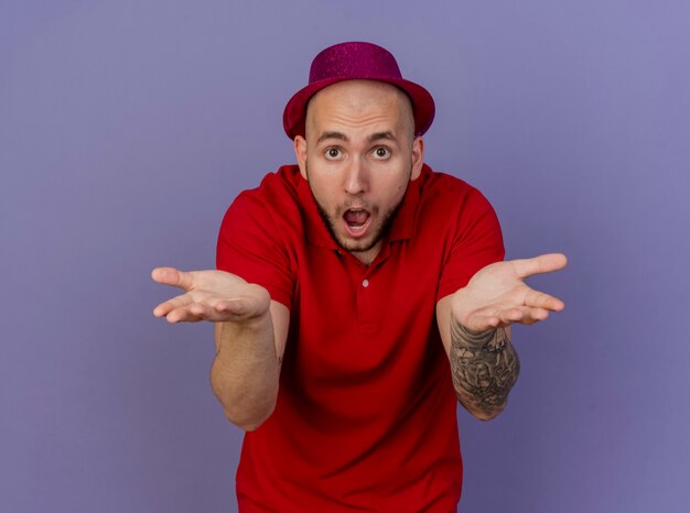 Impressed young handsome slavic party guy wearing party hat looking at camera showing empty hands isolated on purple background