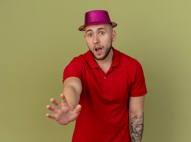 Ragazzo di partito slavo bello colpito giovane che indossa il cappello del partito che guarda l'obbiettivo che allunga la mano verso la macchina fotografica isolata su fondo verde oliva con lo spazio della copia