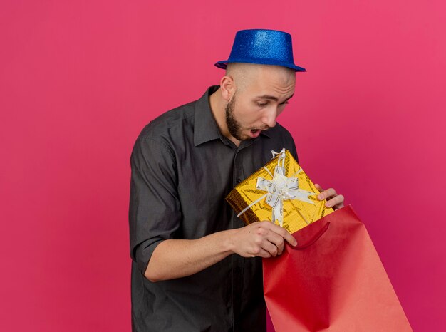 Impressed young handsome slavic party guy wearing party hat pulling out gift pack from paper bag looking at gift pack isolated on crimson background with copy space