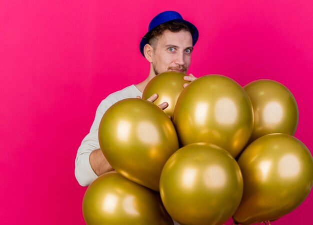 Impressed young handsome slavic party guy wearing party hat standing behind balloons holding one of them looking at camera isolated on crimson background with copy space