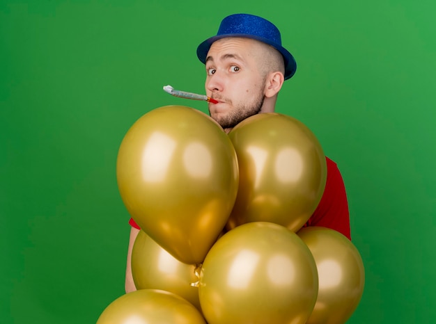 Impressed young handsome slavic party guy wearing party hat standing behind balloons looking at camera blowing party blower isolated on green background with copy space