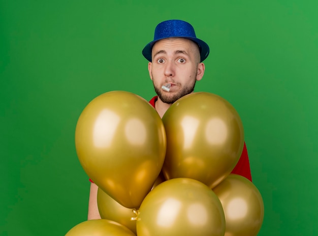 Impressed young handsome slavic party guy wearing party hat standing behind balloons looking at camera blowing party blower isolated on green background with copy space