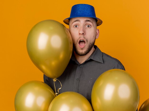 Impressed young handsome slavic party guy wearing party hat standing behind balloons looking at camera isolated on orange background