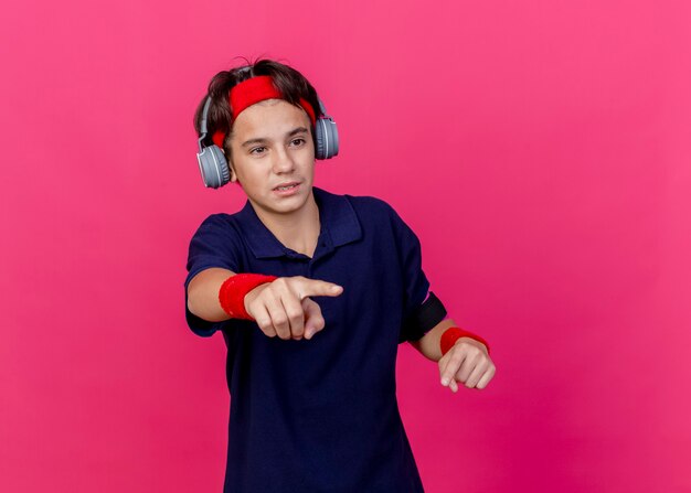 Impressed young handsome sporty boy wearing headband and wristbands and headphones phone armband with dental braces looking and pointing straight isolated on crimson wall with copy space