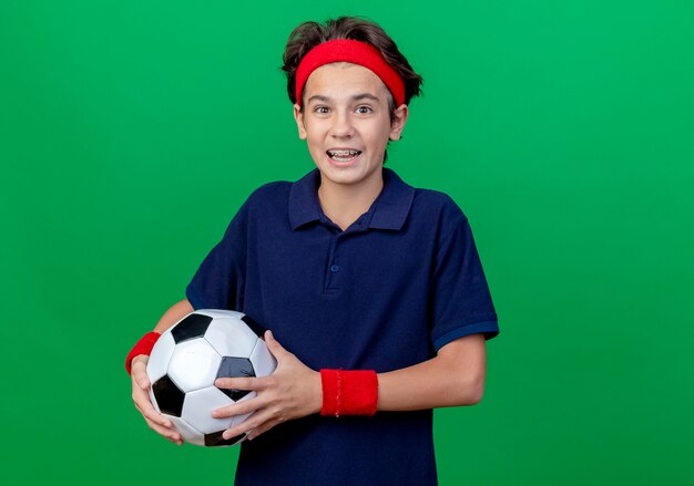 Impressed young handsome sporty boy wearing headband and wristbands with dental braces holding soccer ball looking at camera isolated on green background with copy space