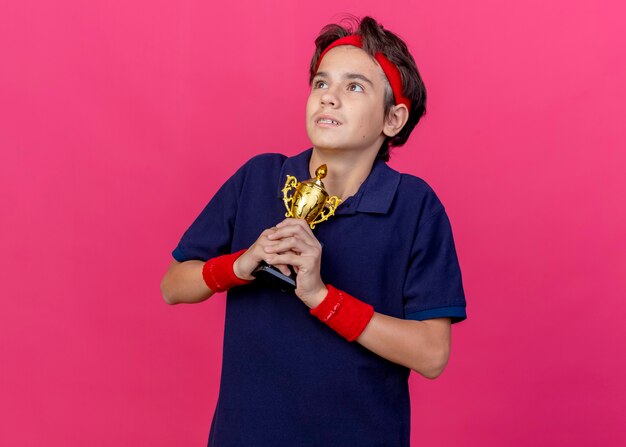 Impressed young handsome sporty boy wearing headband and wristbands with dental braces holding winner cup looking at side isolated on crimson wall with copy space