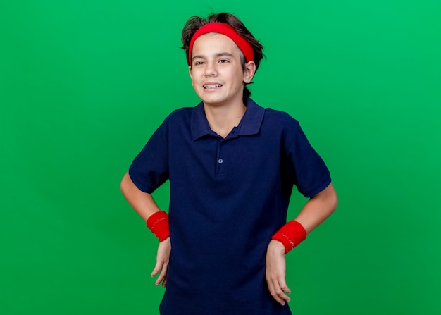 Impressed young handsome sporty boy wearing headband and wristbands with dental braces keeping hands on waist looking straight isolated on green background with copy space