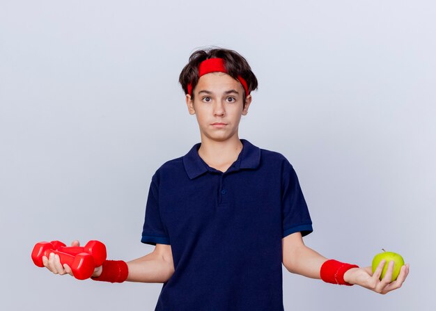 Impressed young handsome sporty boy wearing headband and wristbands with dental braces looking at camera holding dumbbells and apple isolated on white background