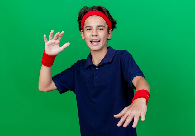 Impressed young handsome sporty boy wearing headband and wristbands with dental braces looking at camera keeping hands in air isolated on green background with copy space