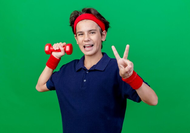 Impressed young handsome sporty boy wearing headband and wristbands with dental braces looking at front doing peace sign isolated on green wall