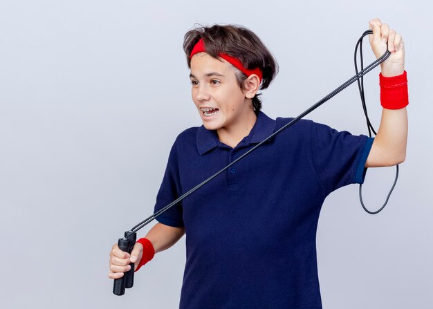 Impressed young handsome sporty boy wearing headband and wristbands with dental braces looking straight holding jump rope isolated on white background with copy space