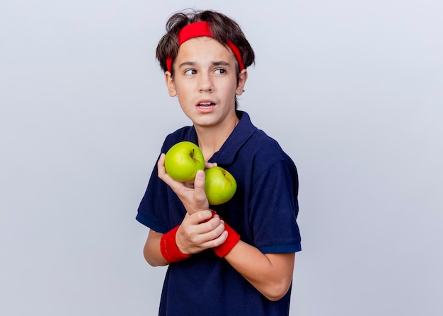 Impressed young handsome sporty boy wearing headband and wristbands with dental braces standing in profile view holding apples and wrist looking at side isolated on white background with copy space