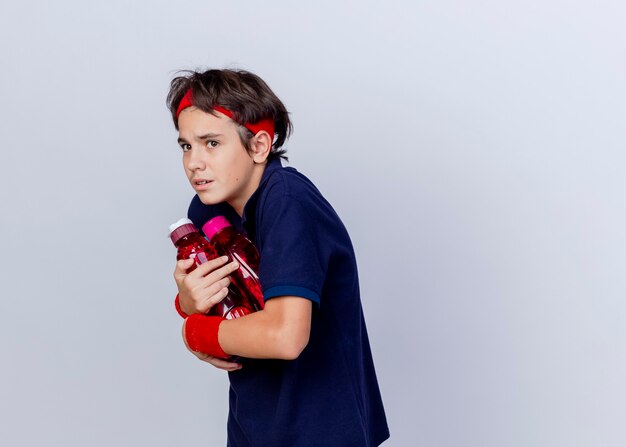 Impressed young handsome sporty boy wearing headband and wristbands with dental braces standing in profile view holding water bottles looking at side isolated on white background with copy space