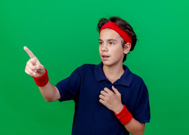 Impressed young handsome sporty boy wearing headband and wristbands with dental braces touching chest looking and pointing straight isolated on green wall