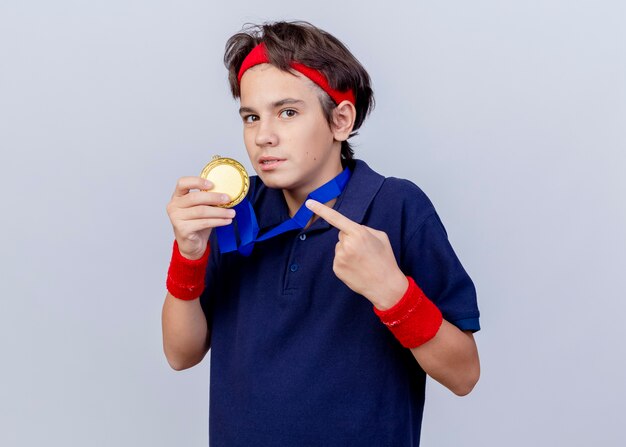 Impressed young handsome sporty boy with medal around neck, holding and pointing at medal