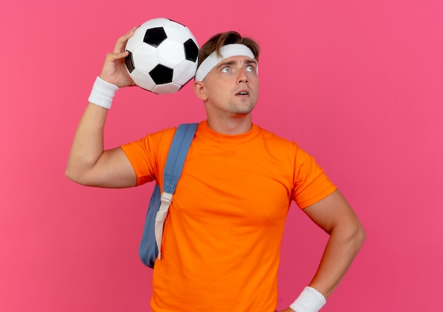 Impressed young handsome sporty man wearing headband and wristbands and back bag holding soccer ball and touching head with it looking up with hand on waist isolated on pink 