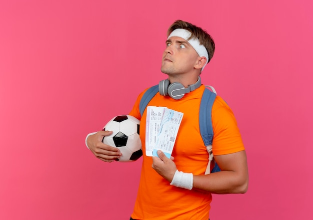Впечатленный молодой красивый спортивный мужчина с повязкой на голову, браслетами и задней сумкой с наушниками на шее, держащий билеты на самолет и футбольный мяч, выглядящий прямо