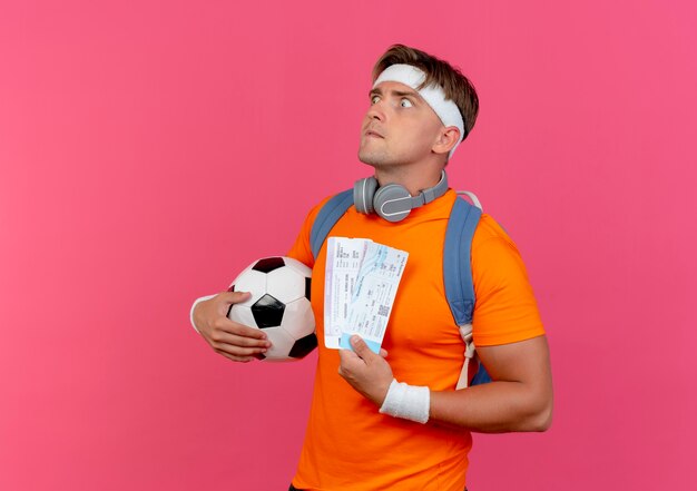 Impressed young handsome sporty man wearing headband and wristbands and back bag with headphones on neck holding airplane tickets and soccer ball looking straight