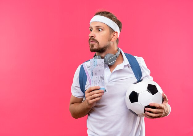 Impressed young handsome sporty man wearing headband and wristbands and back bag with headphones on neck