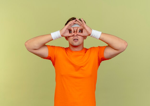 Impressed young handsome sporty man wearing headband and wristbands doing look gesture isolated on olive green