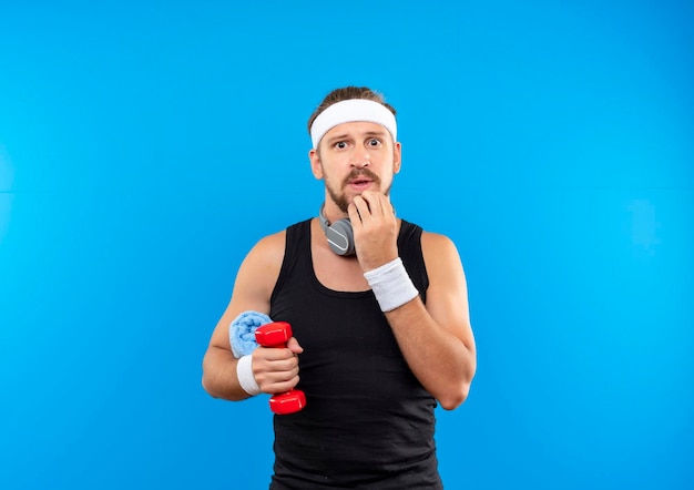 Impressed young handsome sporty man wearing headband and wristbands and headphones around neck holding dumbbell with towel and putting hand on chin isolated on blue wall with copy space