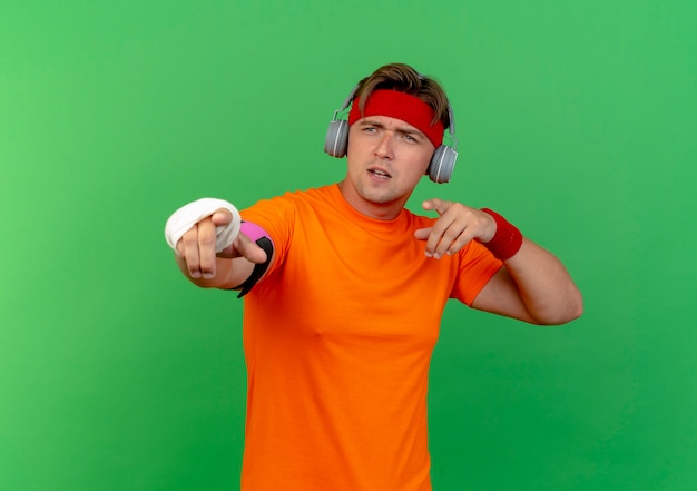 Free photo impressed young handsome sporty man wearing headband and wristbands and headphones and phone armband with injured wrist wrapped with bandage pointing and looking at side isolated on green