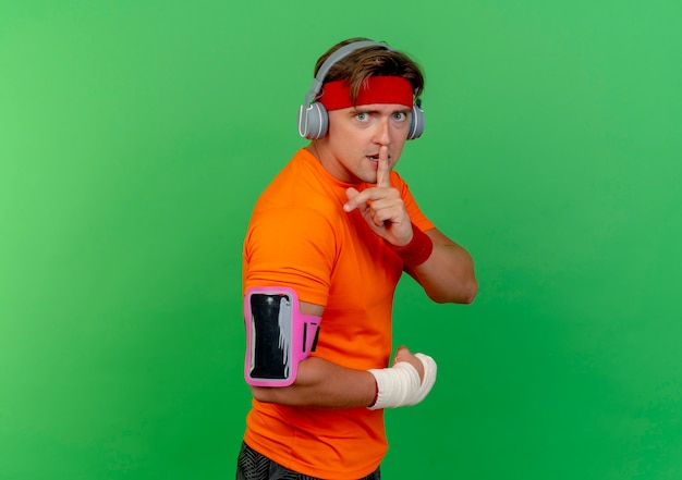 Impressed young handsome sporty man wearing headband and wristbands and headphones and phone armband with wrist wrapped with bandage standing in profile view gesturing silence