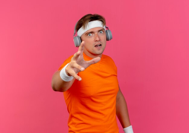 Impressed young handsome sporty man wearing headband and wristbands and headphones stretching out hand isolated on pink