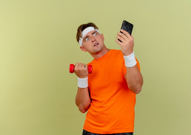 Impressed young handsome sporty man wearing headband and wristbands holding mobile phone and dumbbell looking at phone isolated on olive green