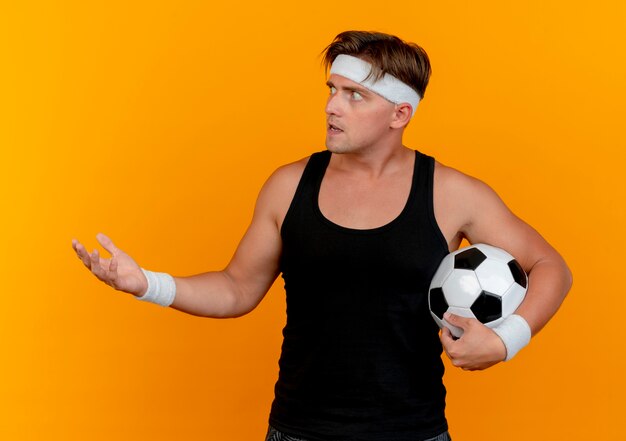 Impressed young handsome sporty man wearing headband and wristbands holding soccer ball looking at side and showing empty hand isolated on orange