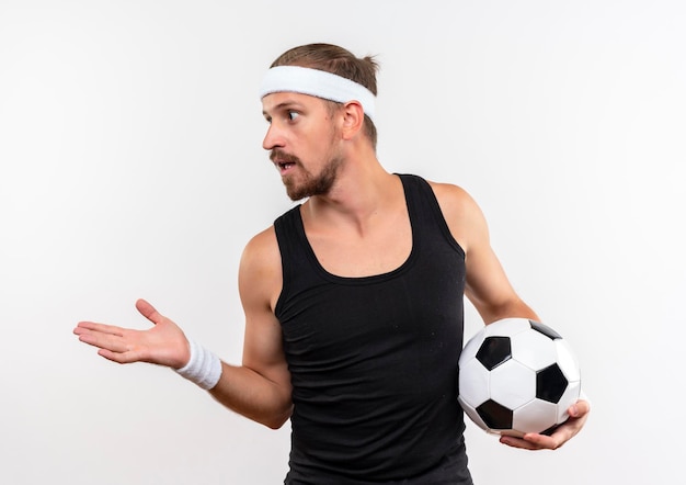 Impressed young handsome sporty man wearing headband and wristbands holding soccer ball looking at side showing empty hand isolated on white wall