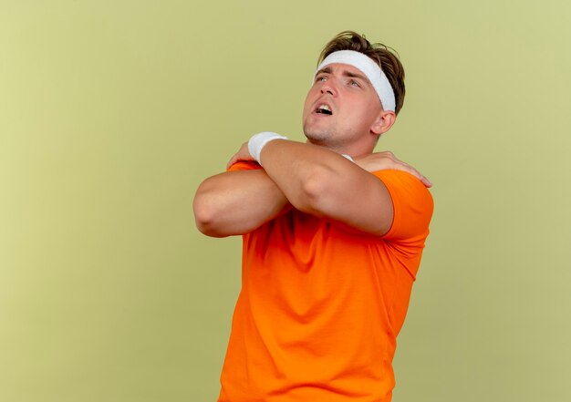 Impressed young handsome sporty man wearing headband and wristbands looking up putting hands on shoulders isolated on olive green