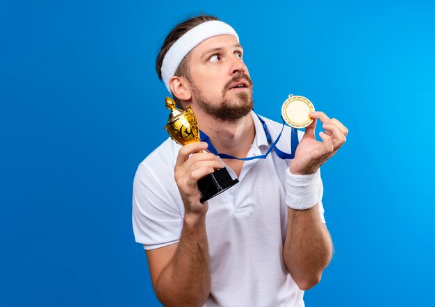 Impressed young handsome sporty man wearing headband and wristbands and medal around neck holding medal and winner cup looking at side isolated on blue wall with copy space